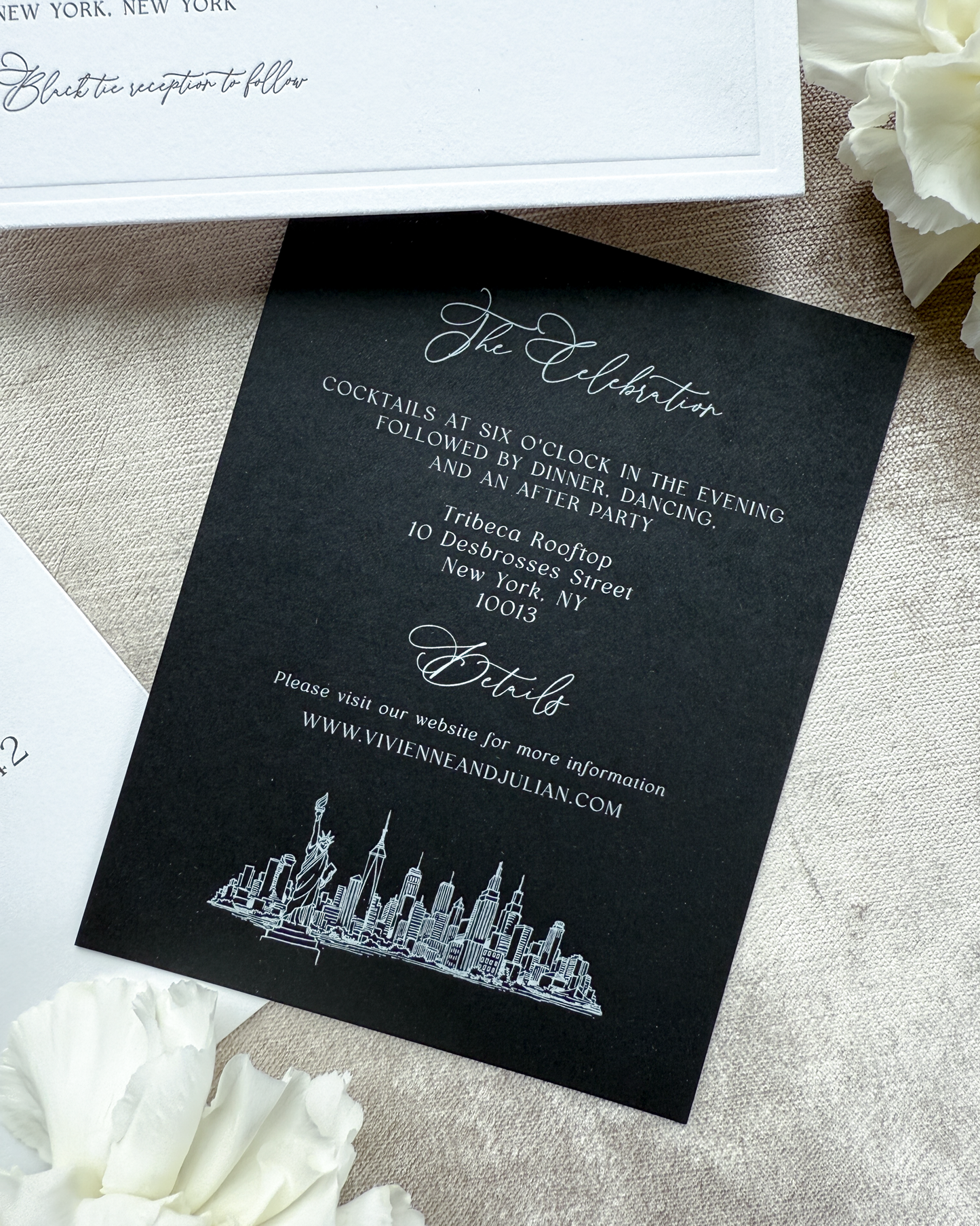 Black invitation card with white text and skyline design on a textured surface with white flowers.